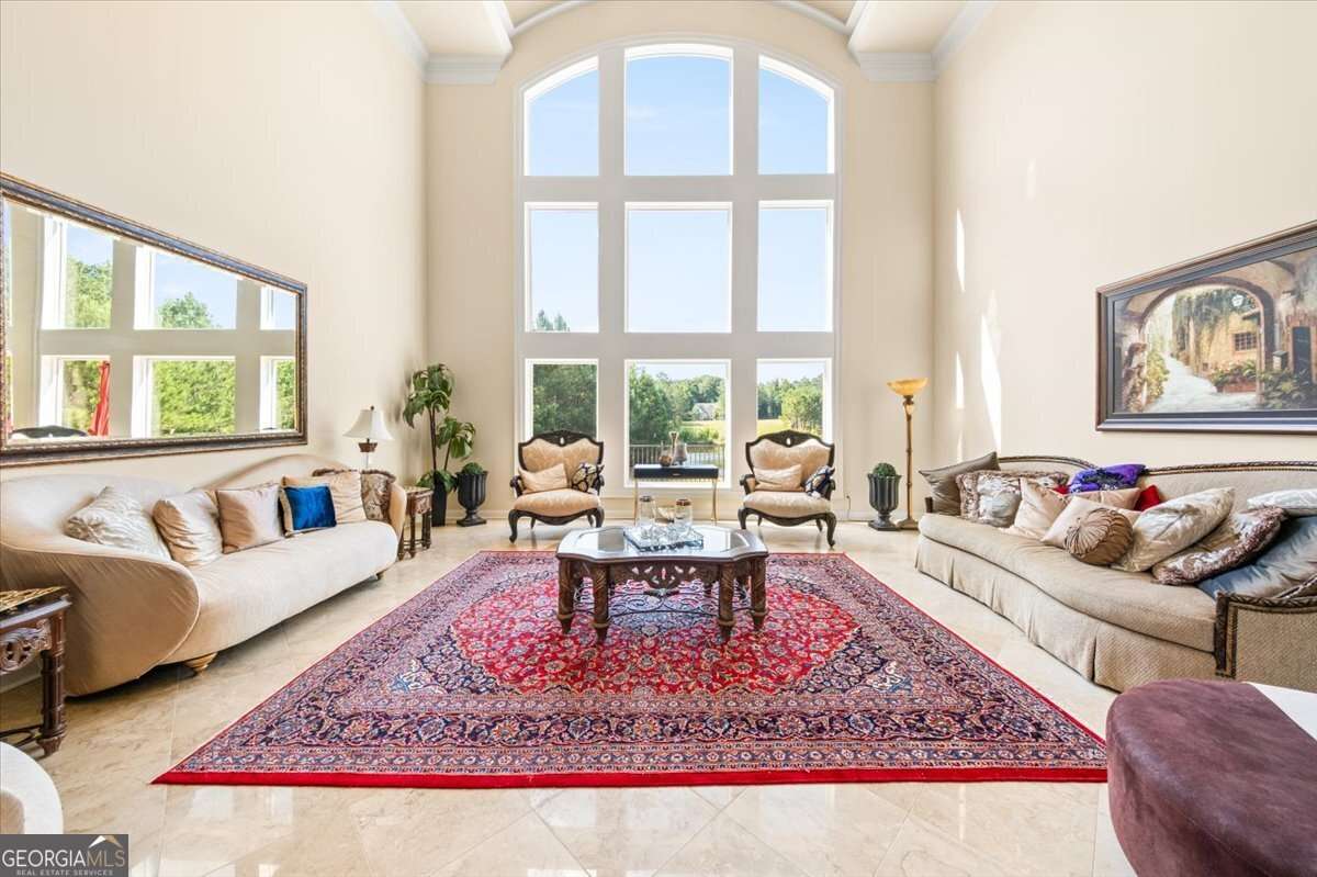 Bright living room with a tall arched window, two beige sofas, matching chairs, and a red patterned rug in the center.