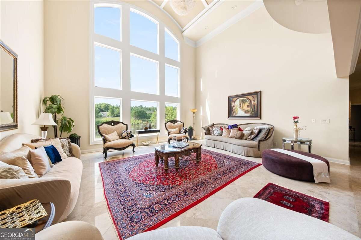 Sunlit living room with tall arched window wall, cream sofas, and ornate chairs around a wood coffee table on a red Persian rug.