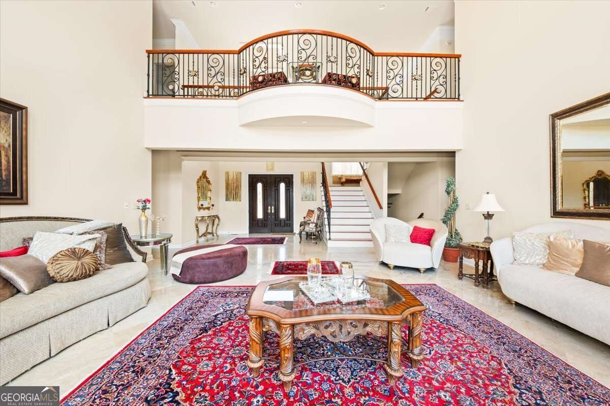 Luxurious two-story living room with ornate iron balcony, beige sofas, and a carved wooden coffee table on a red Persian rug.Elegant furniture and a staircase create a grand entry hall feel.