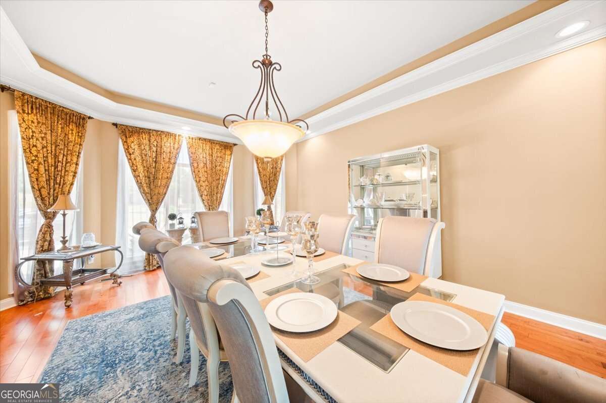 Elegant dining room with a glass-top table set for six, beige upholstered chairs, chandelier, and gold-patterned drapes over large windows.