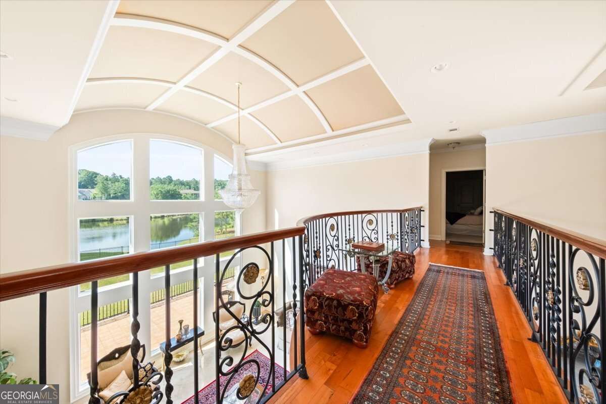 Upscale upstairs hallway with decorative wrought-iron railing, a long patterned rug, and a crystal chandelier overlooking a lake view through large arched windows. A bedroom doorway is at the far end.