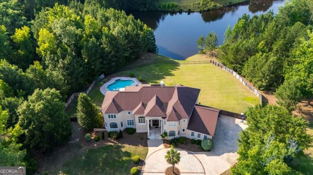 Aerial view of a large beige house with brown roof, circular driveway, and a kidney-shaped pool, surrounded by a fenced yard and trees near a lake; wooded backdrop.