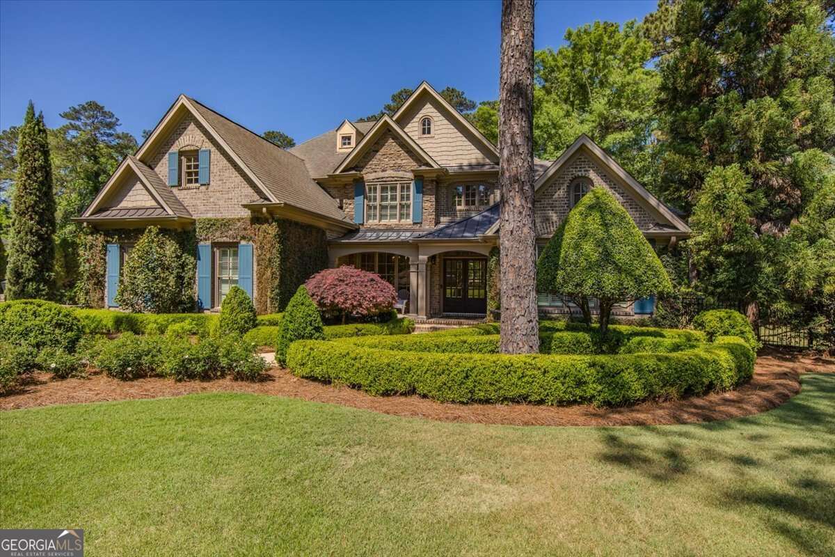 Front view of a large brick-and-stone suburban house with blue shutters, surrounded by manicured hedges and trees.