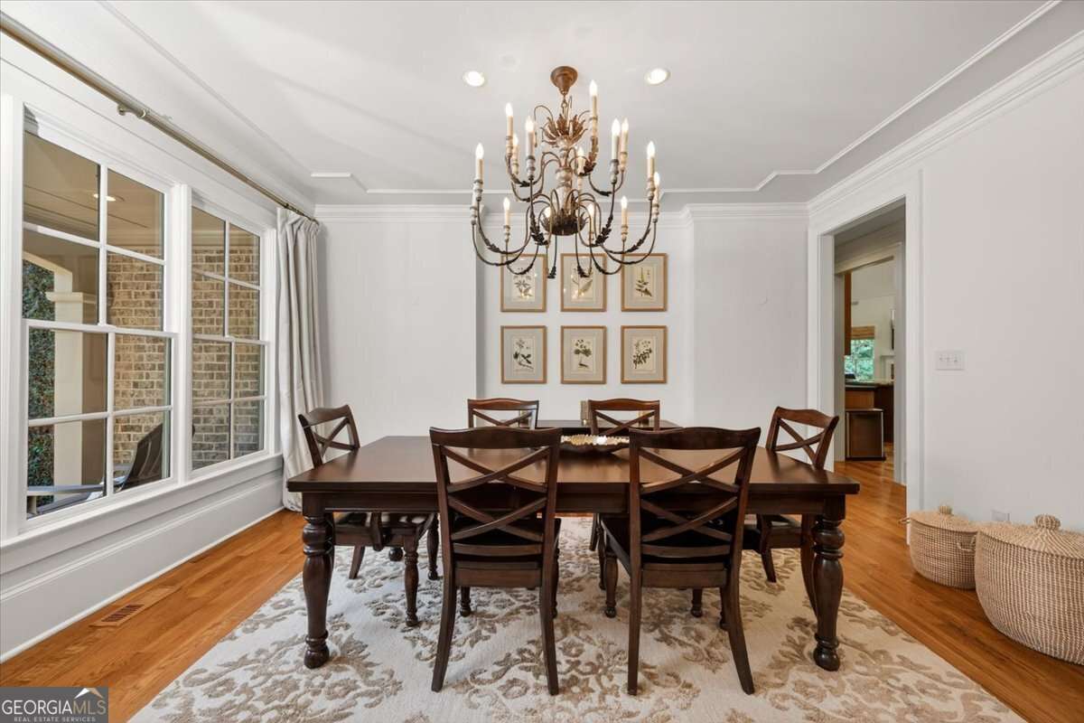 Formal dining room with a dark wood table, six matching chairs, and a wrought-iron chandelier above; framed botanical prints on the wall, light walls, and a large window with curtains.