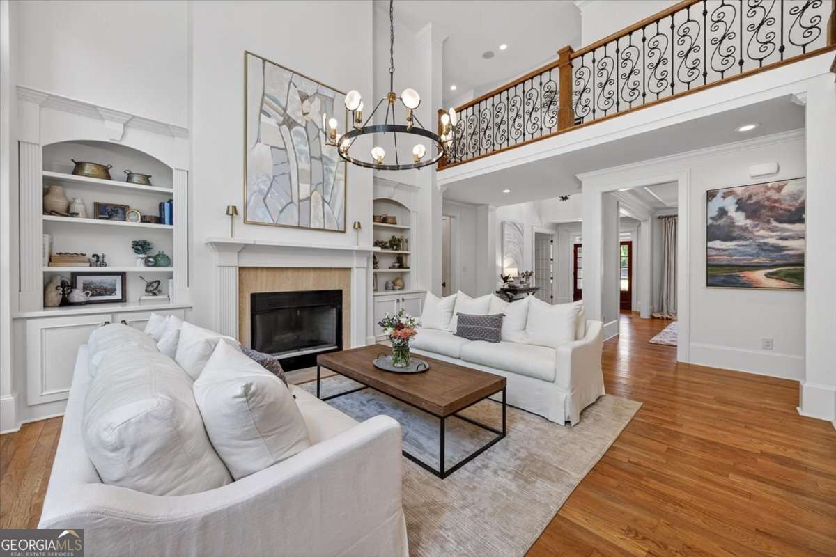 Bright two-story living room with white sofas, a fireplace, built-in shelves, abstract art above the mantel, warm wood floors, and a wrought-iron balcony railing.