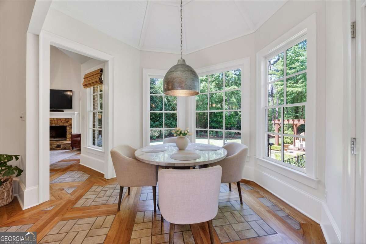Bright dining nook with a round glass table and four beige upholstered chairs set beside large white-framed windows overlooking a green backyard.