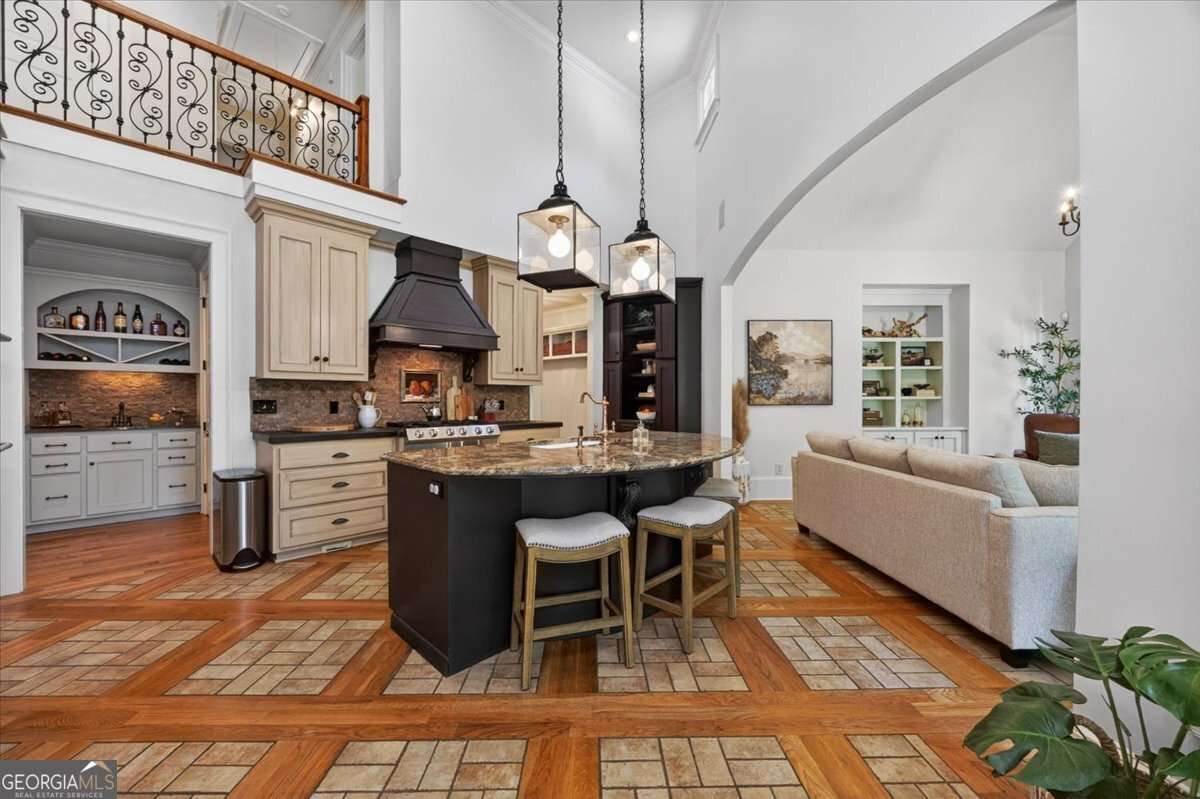 Open-concept kitchen with a large granite island, beige cabinets, a black range hood, and two hanging lantern pendant lights.