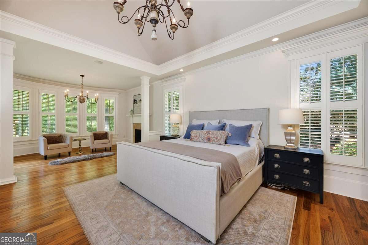 Bright luxury bedroom with a white upholstered king bed, navy nightstands, chandelier, and a seating area by large windows with shutters.