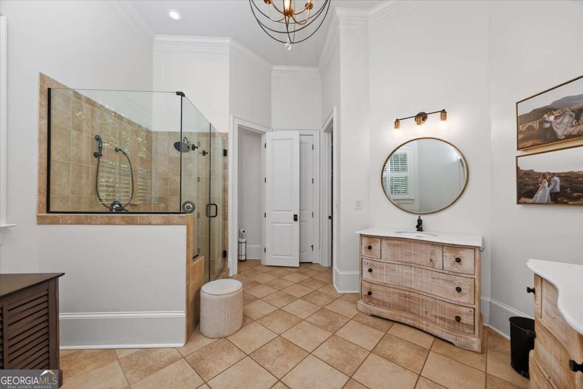 Bright bathroom with glass-walled shower, beige tiles, and a rustic wood vanity with a round mirror above. Two landscape photos on the wall.