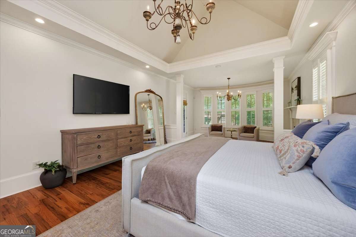 Bright master bedroom with a wall-mounted TV above a wooden dresser, a beige upholstered bed, and a bay-window seating area beyond the foot of the bed.