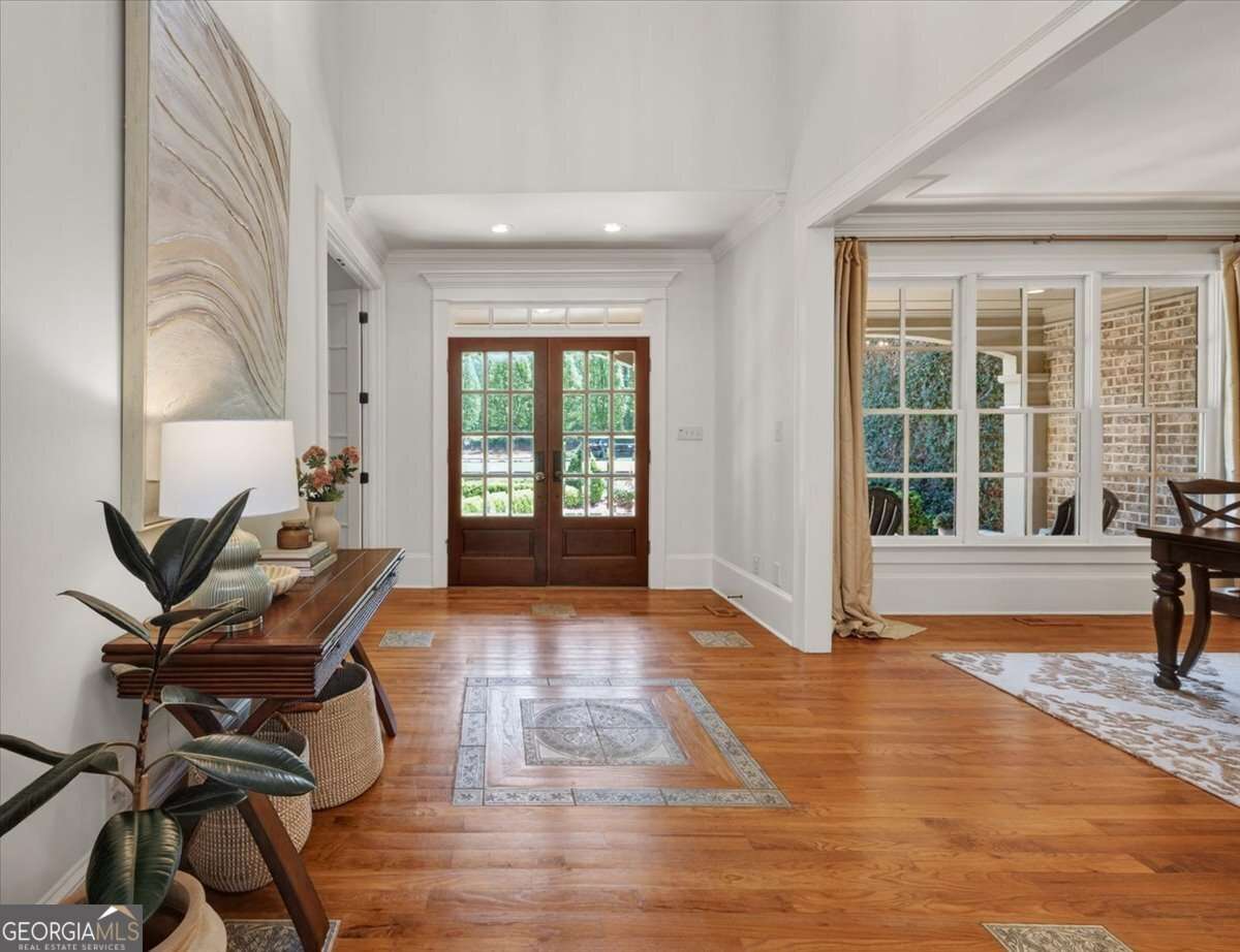 Bright entry hall with hardwood floors, glass-pane front doors, and a console table with lamp on the left; dining area visible to the right.