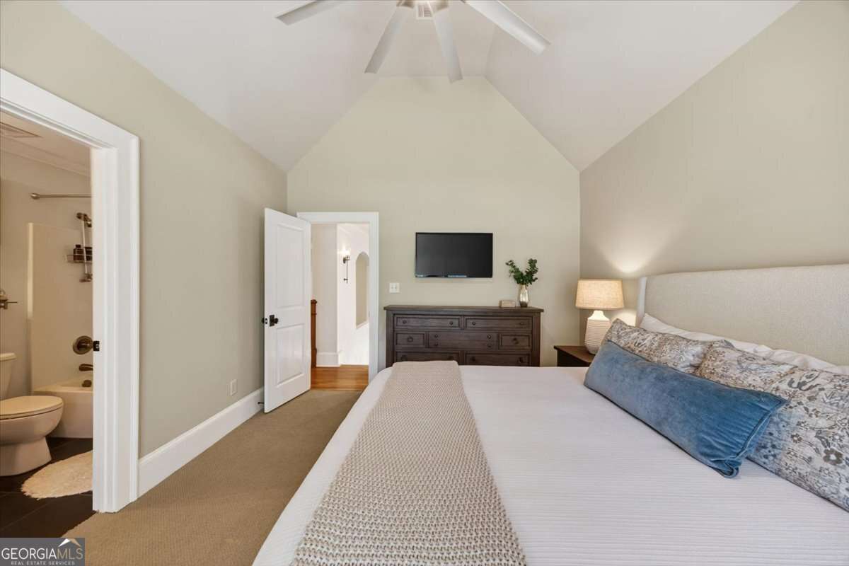 Bedroom with a large bed, beige decor, and a dresser with a wall-mounted TV across from the bed; open doorway to a bathroom on the left.