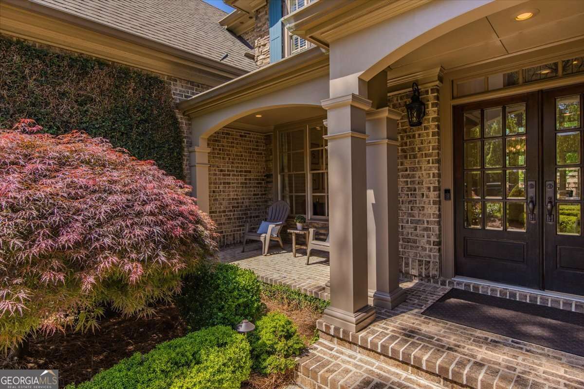 Front porch of a brick house with two chairs, small table, and a potted plant under a covered entrance. Lush bushes and a Japanese maple frame the scene.