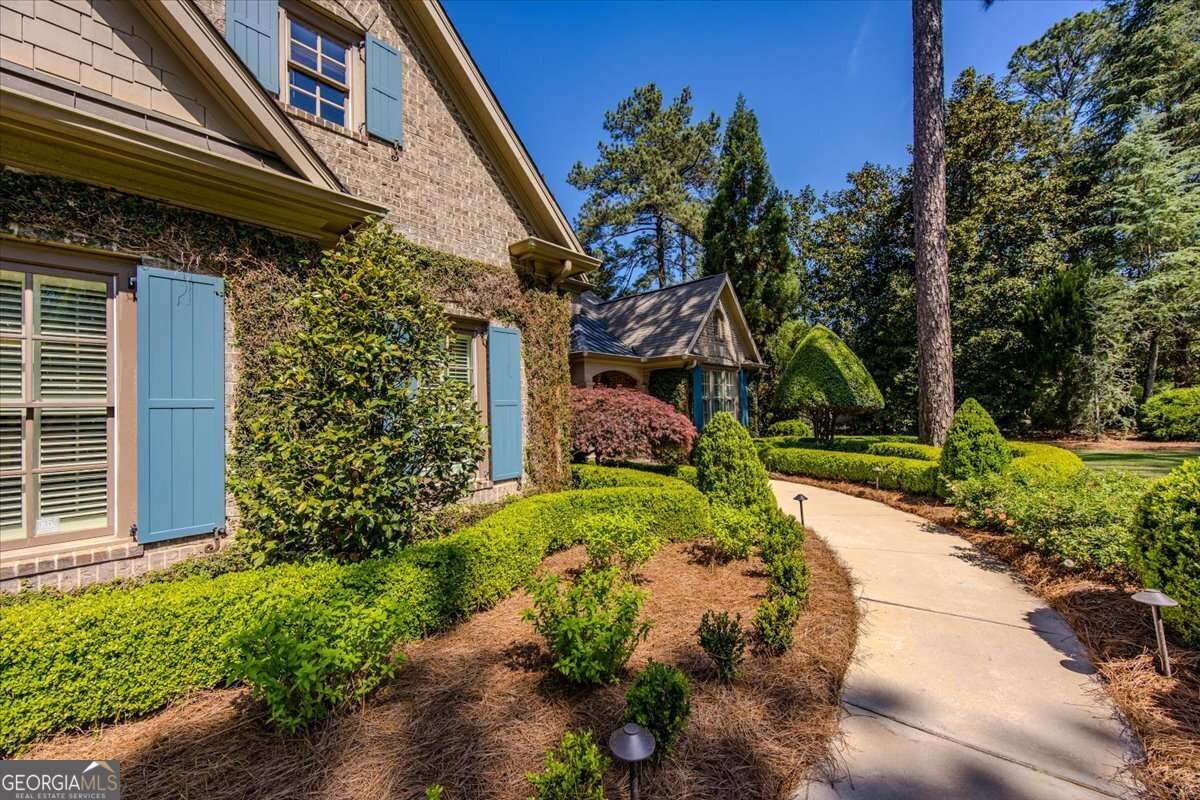 Front yard of a brick house with blue shutters, trimmed hedges, and a curved concrete walkway under a bright blue sky.