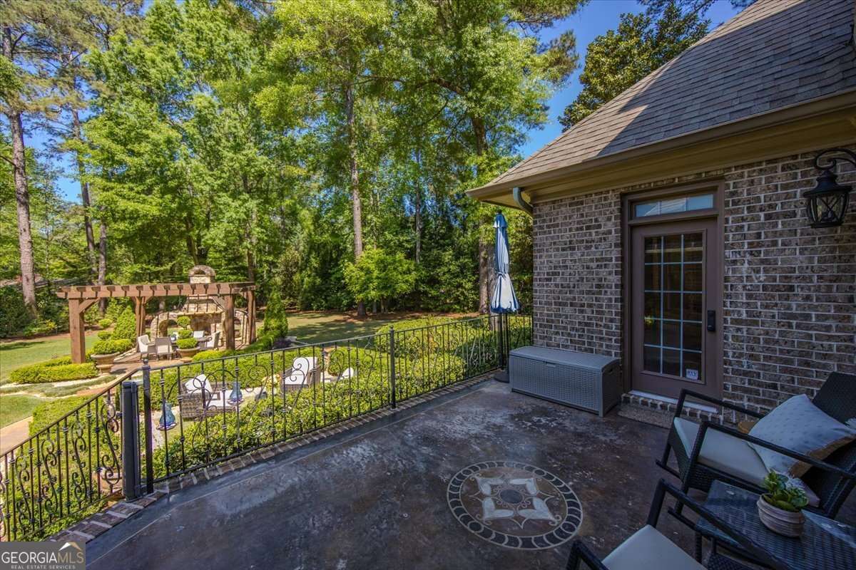 Brick house patio with wrought-iron railing, outdoor seating, and a circular tile star motif on the concrete overlooking a lush backyard with a pergola.