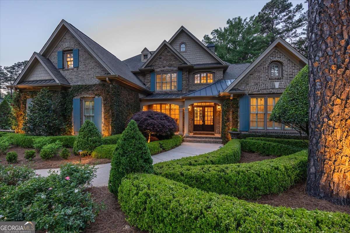 Front view of a large brick-and-stone suburban house with blue shutters, surrounded by manicured hedges and trees.