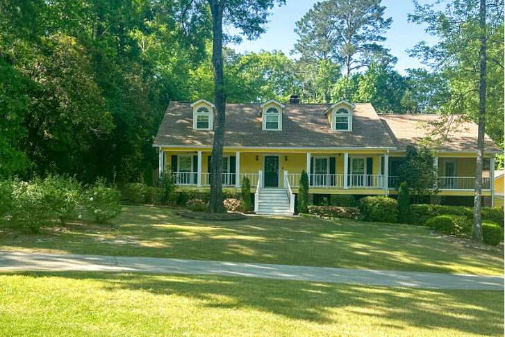 Yellow ranch-style house with a wraparound porch, three dormer windows, and a well-kept front yard.