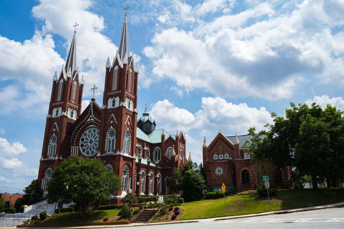 Red brick Gothic church with twin spires and a large rose window, set against a blue sky with clouds and green landscaping nearby.