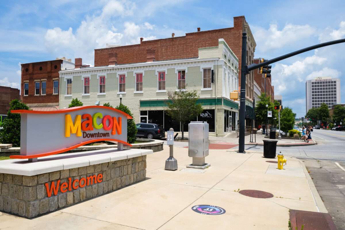 Colorful 'Macon Downtown' sign in the foreground with brick and beige storefronts along a sunny city street in Macon.