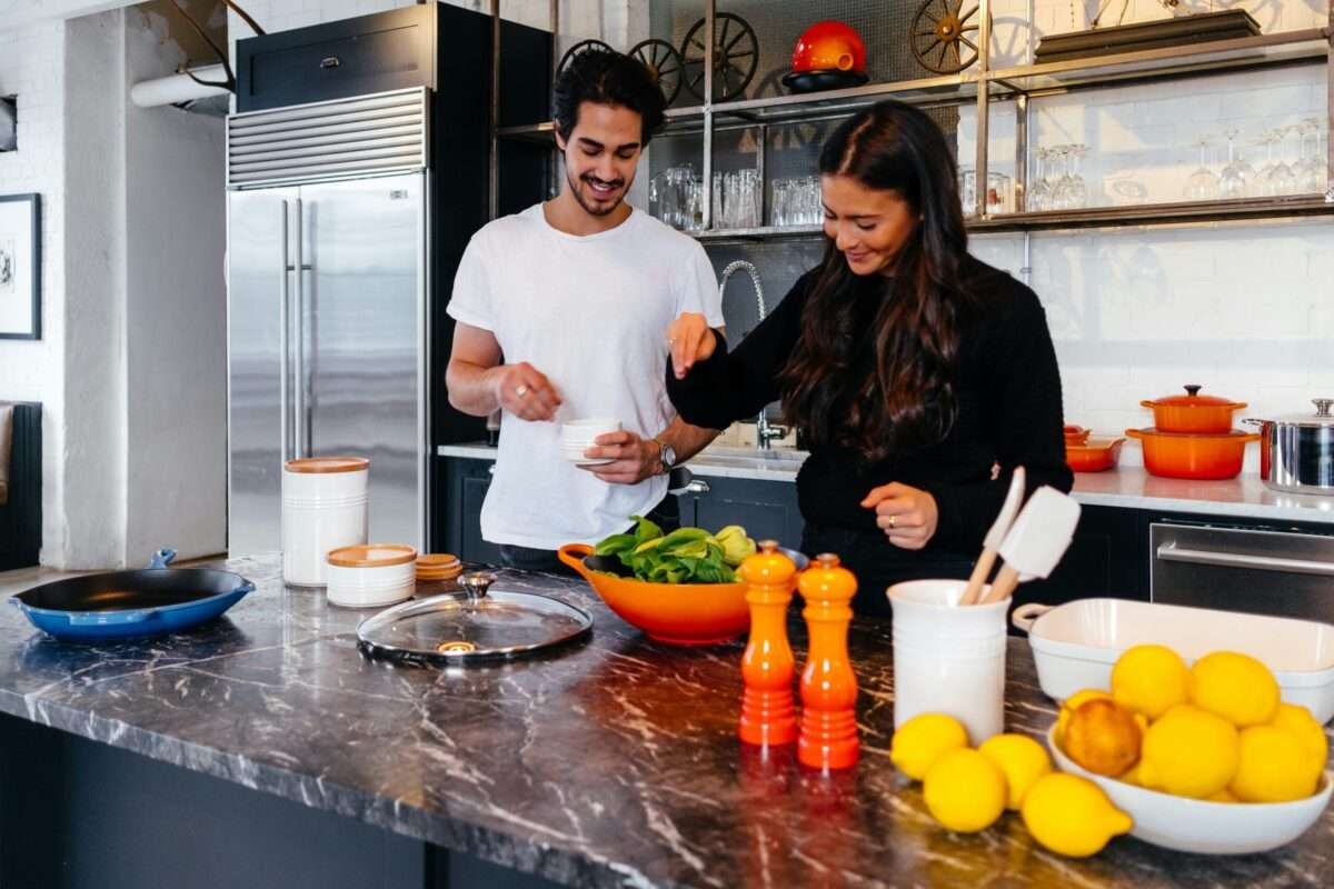 Couple cooking together at a marble kitchen island, adding ingredients to a bowl with greens and lemons nearby.