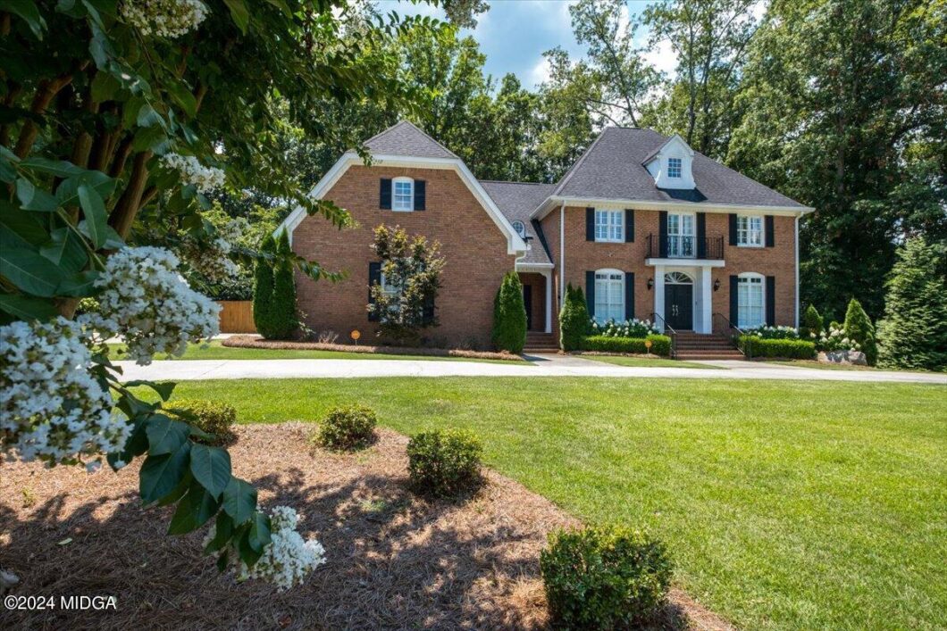 Brick two-story brick colonial-style house with black shutters, white trim, and a small balcony, set in a manicured yard with trees and shrubs around.