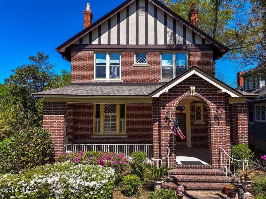Brick two-story house with arched porch, American flag, and blooming garden in front yard.