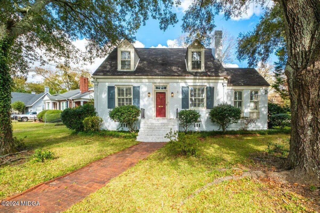 Front view of a white cottage with blue shutters, a red door, and a brick walkway lined by shrubs.