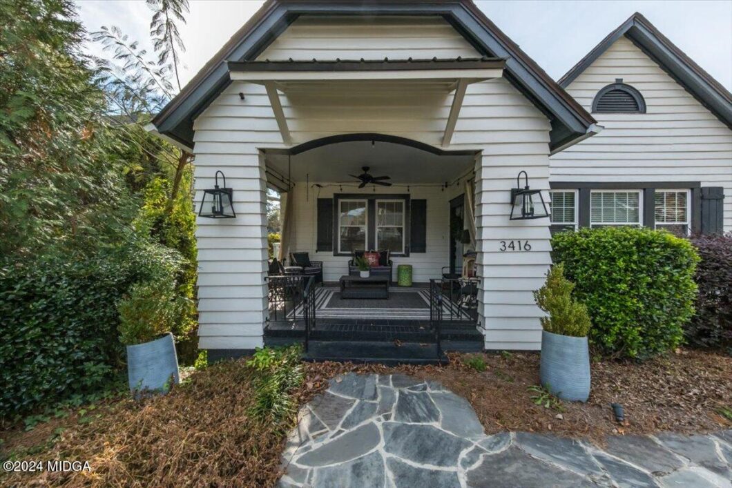 White front porch with seating area under a peaked roof, address 3416, stone walkway leading to steps, surrounding shrubs and lanterns.