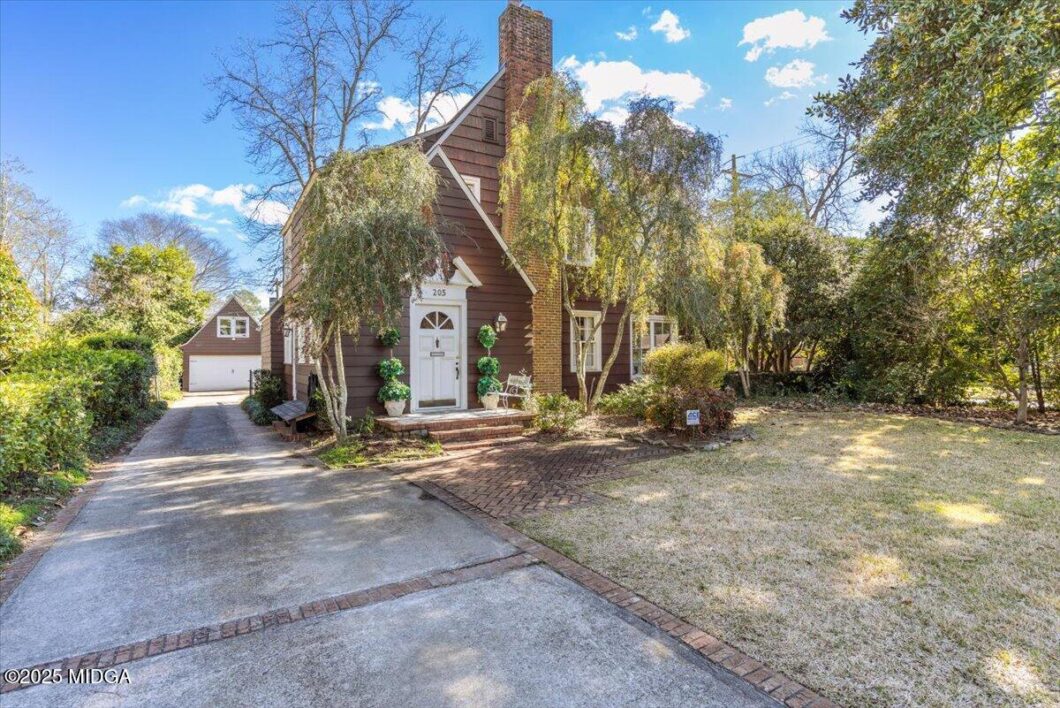 Charming brown A-frame house with a white front door, brick steps, and a brick-paved path surrounded by trees on a sunny day.