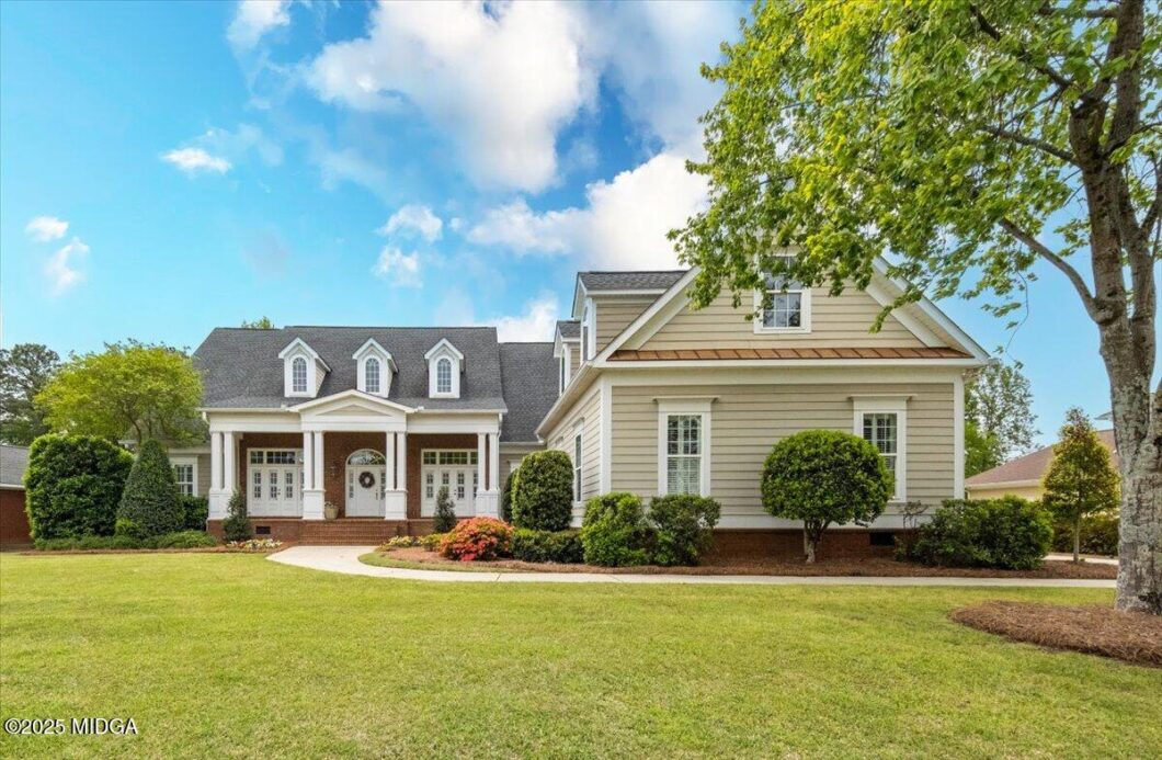 Large suburban house with beige siding, white trim, brick front porch, and three dormer windows under a gray gabled roof; well-kept lawn and curved walkway lead to the front door on a sunny day