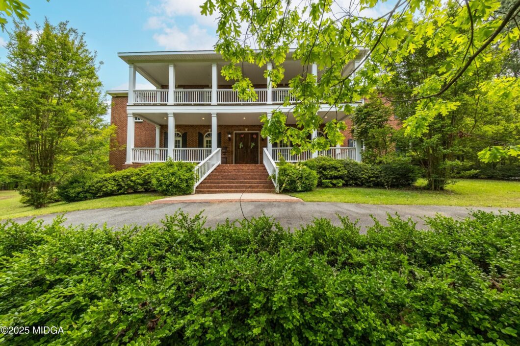 Brick two-story house with white-columned porch, brick steps, and lush green hedges and trees surrounding it.