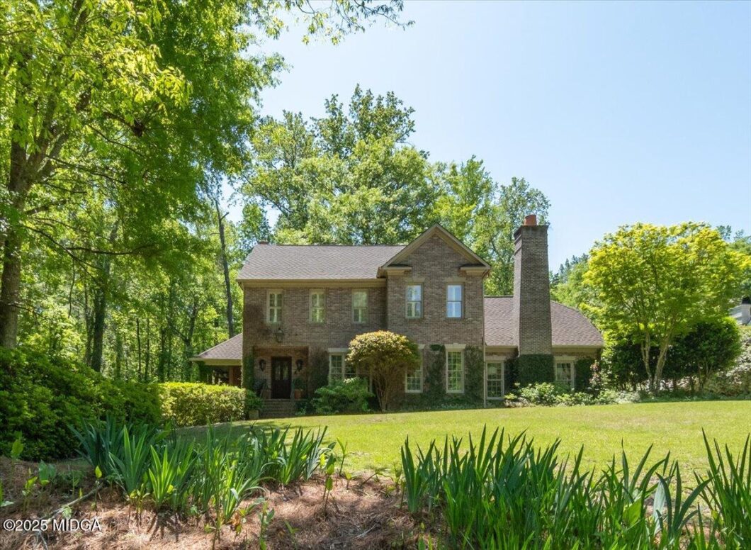 Brick two-story house with central gable, large chimney, and a green lawn surrounded by trees on a sunny day from the yard edge.