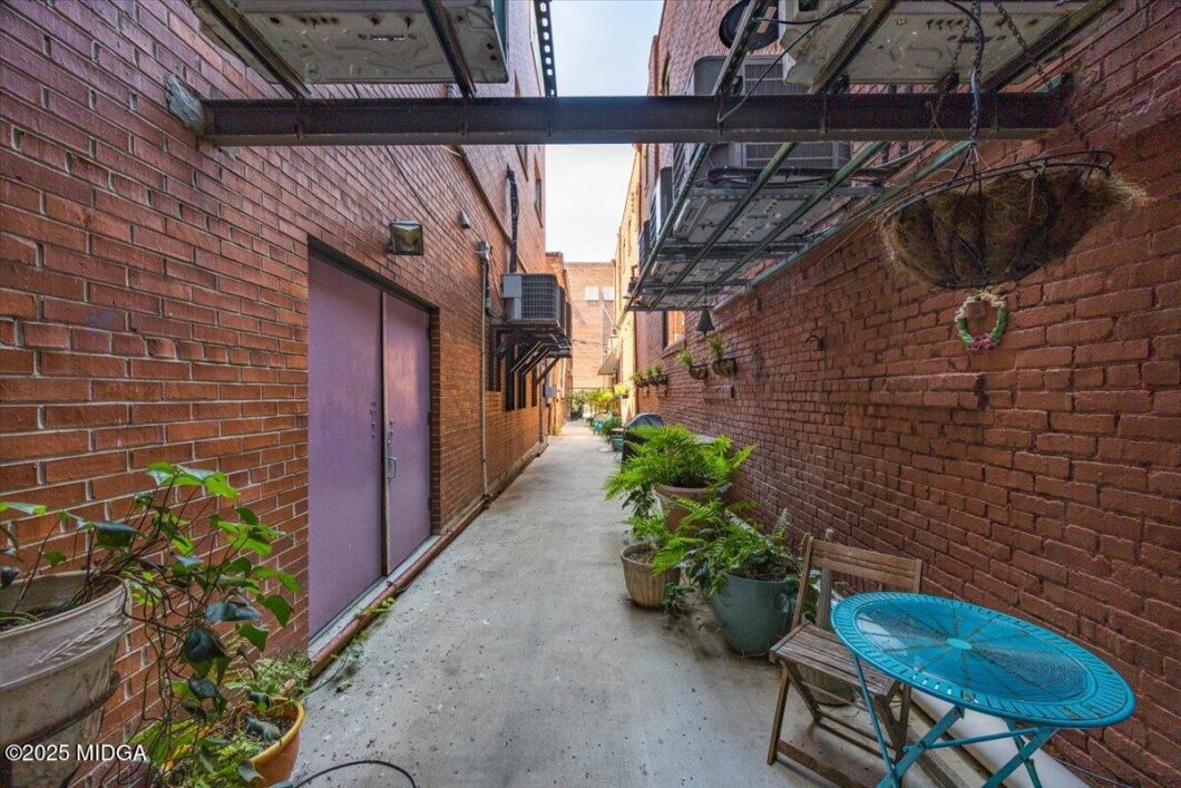 Narrow brick alley with a purple door, potted plants along the wall, and a blue round table with a chair on the right side.