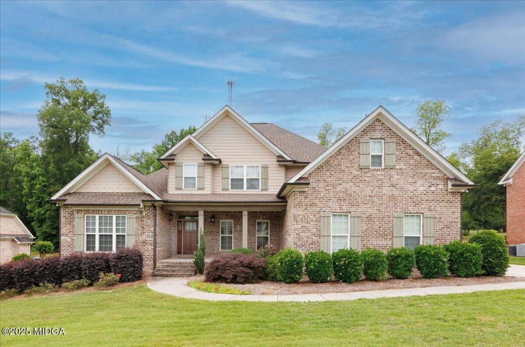 Two-story brick house with beige siding, front porch, and neatly trimmed shrubs in a suburban yard.