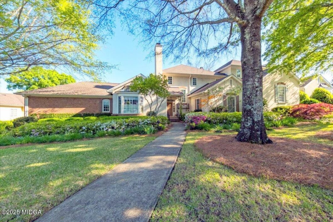 Front view of a suburban house with a manicured lawn and a concrete walkway leading to the front door, large tree in the yard and flowering shrubs.
