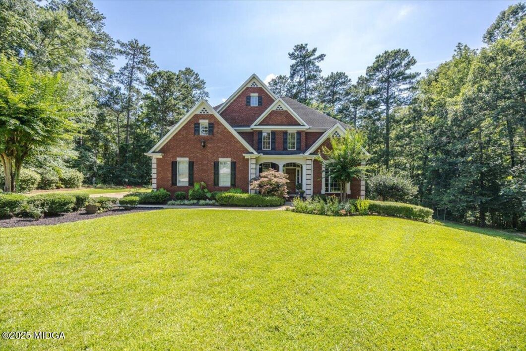 Two-story brick house with white trim and a covered front porch, set on a large manicured lawn with mature trees nearby.