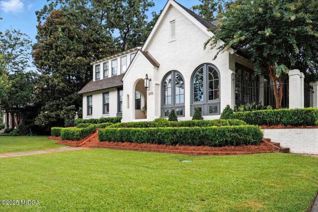 White brick house with large arched windows and a covered entry, framed by manicured hedges and a green lawn on display edge.