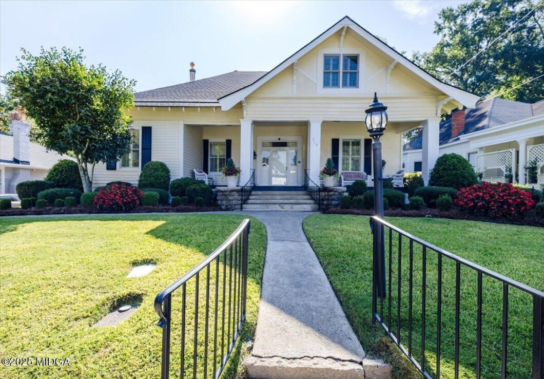 White bungalow with a front porch, black railings, symmetrical landscaping, and a curved concrete path to the entrance.