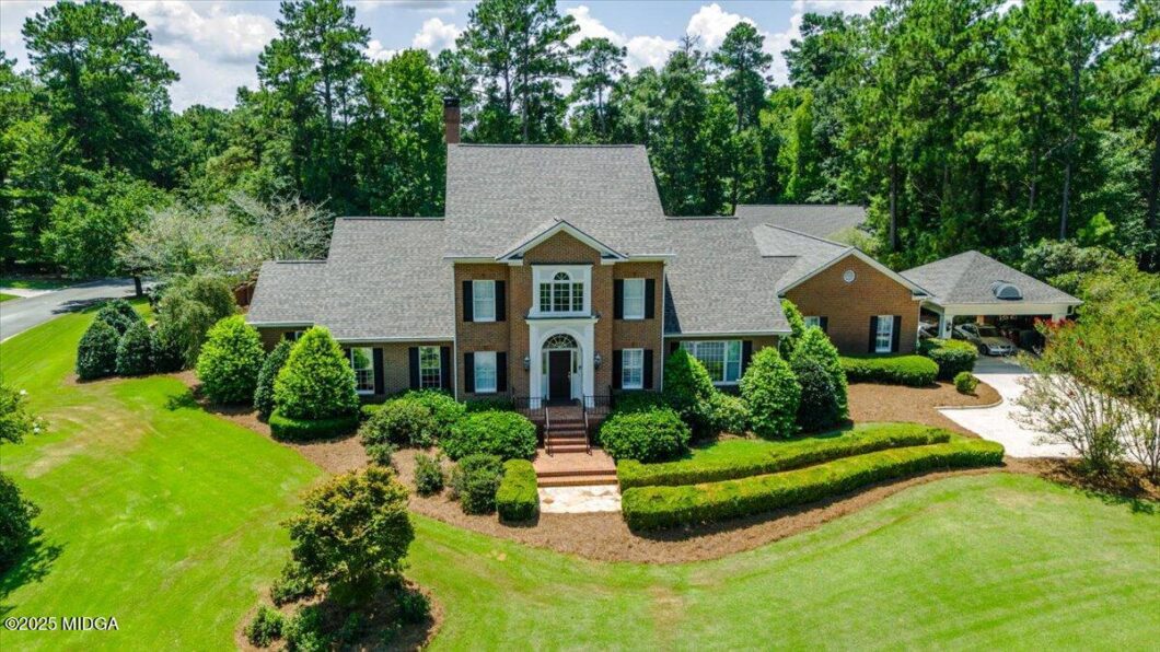 Aerial view of a brick two-story house with a steep gray roof, surrounded by manicured lawn and tall trees.