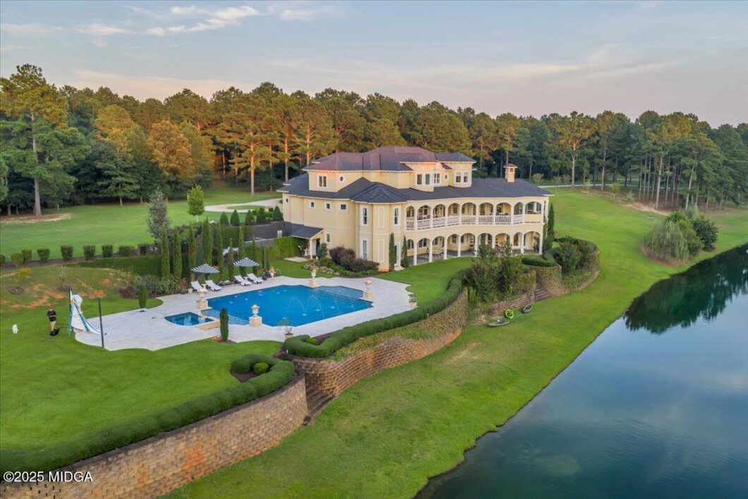 Aerial view of a large cream-colored mansion with a tiled roof, curved balcony, and surrounding pool on a manicured lawn beside a lake.