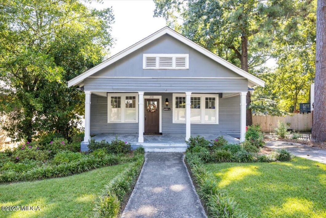 Blue-gray bungalow with a covered front porch supported by white columns, walkway leading to a dark front door with a wreath, surrounded by a manicured lawn and shrubs.