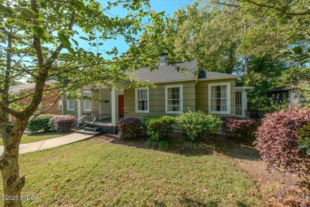 Front view of a single-story beige house with a small porch, red door, and white window frames, surrounded by shrubs and trees.