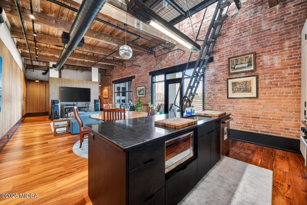 Industrial loft interior with brick walls, exposed ductwork, and a black island kitchen opening to a living area with wooden floors.