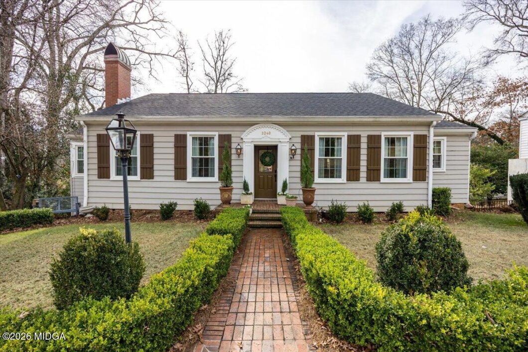 A single-story beige house with brown shutters, brick walkway, and symmetrical potted evergreens leading to a front door with a wreath.