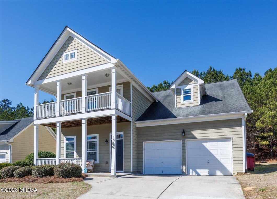 Front view of a beige two-story house with white wraparound porch and two-car garage under a clear blue sky, with manicured shrubs.