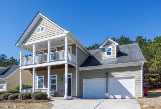 Front view of a beige two-story house with white wraparound porch and two-car garage under a clear blue sky, with manicured shrubs.