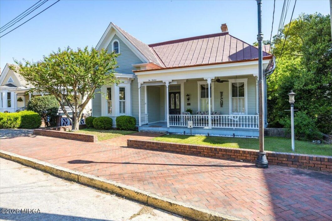 Pastel blue Craftsman-style house with white porch railing, red metal roof, and a brick-paved sidewalk in front.
