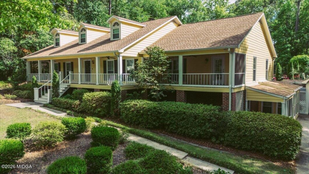 Two-story yellow house with a wraparound porch surrounded by manicured hedges and trees in a suburban yard