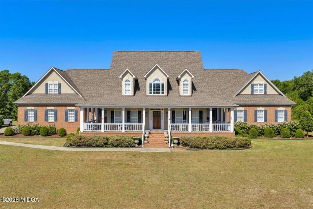 Front view of a large brick house with a wraparound porch, central steps, and dormer windows under a bright blue sky.