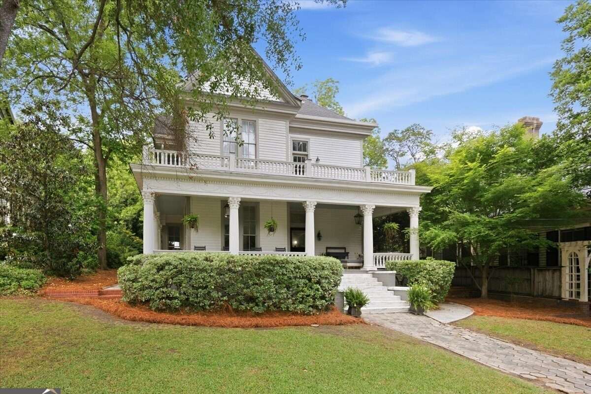 Stately two-story historic home with wraparound porch, tall columns, upper balcony, manicured landscaping, and mature trees on a spacious lot.